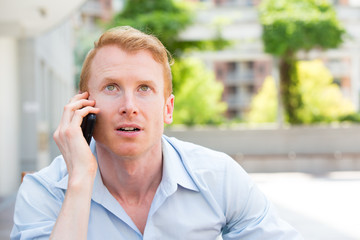 closeup portrait, young man perplexed, baffled, befuddled, bewildered by someone talking on his mobile phone, isolated outdoors background