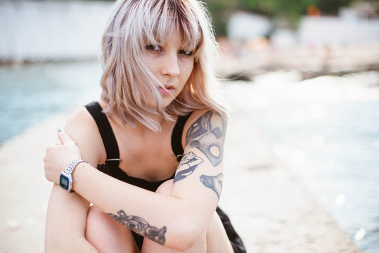 Close up portrait of young hipster girl with stylish blond hair, piercing, tattoos in black dress sitting on the big stone near the sea