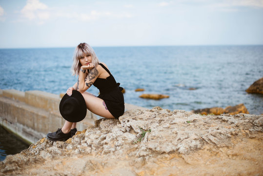 Beautiful young blonde hipster girl with tattoos wearing black dress, holding black hat sitting on the stony beach near the sea. Outdoors