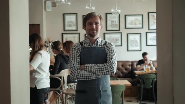 Portrait of smiling owner standing at his restaurant gate. Young entrepreneur leaning at the cafeteria door and looking at camera. Chef or waiter standing in front of coffee shop.