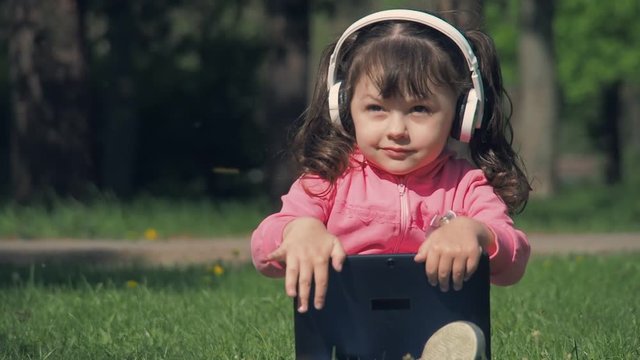 Happy child with tablet in the park. Little girl with headphones and tablet on green grass.