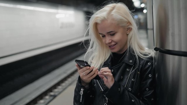 Young blonde travel woman holding a cell phone in her hand waiting on the platform of a railway train. Public transport.
