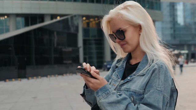 Beautiful young caucasian woman looking at her smartphone and smiling in urban background. Blond girl wearing casual clothes sitting on stairs.