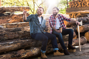 Fototapeta premium smiling lumberjack with coffee cup pointing by finger to partner with axe sitting on logs at sawmill