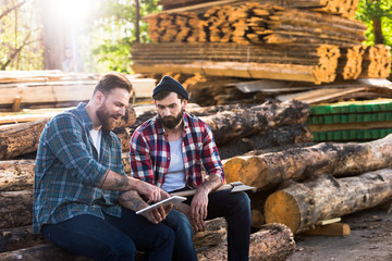 bearded lumberjacks sitting on logs with axe and digital tablet at sawmill