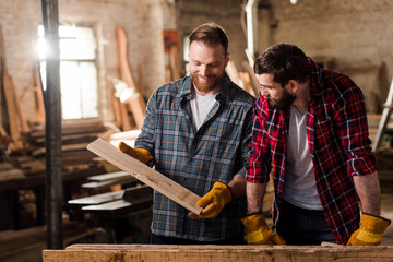 happy carpenter in protective gloves showing wooden plank to partner at sawmill - obrazy, fototapety, plakaty