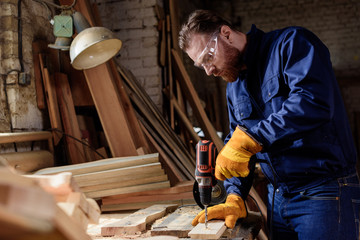 bearded carpenter in protective gloves and googles using electric drill at sawmill