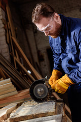 bearded craftsman in protective googles and gloves using grinding machine at sawmill