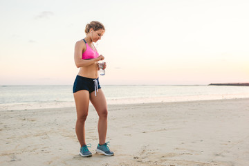 attractive sportswoman in earphones with smartphone in armband case holding bottle of water on beach with sea behind