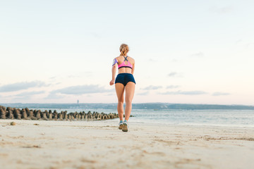 rear view of sportswoman with smartphone in running armband case jogging on sandy beach