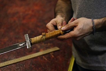 Smith makes leather winding of sword. Man is working in workshop. He fixes leather band on hilt of sword.