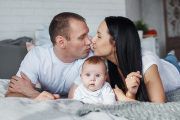 Fototapeta premium Young mom and dad and their baby son lie in bed and look at the camera indoors. The concept of a happy family