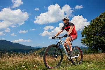 Fototapeta na wymiar Professional sportsman rider in sportswear and helmet cycling bicycle in high grass, male cyclist enjoying view of Carpathian mountains on sunny summer day. Active lifestyle and extreme sport concept