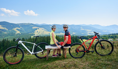 Happy couple sportsman cyclists sitting on wooden bench near bicycles, looking to the camera. Tourists resting after riding on bikes in mountains. Active lifestyle, traveling and outdoor sport concept