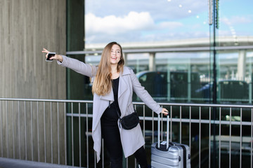 European female person stopping taxi with raised hand, valise and smartpone near airport. Concept...