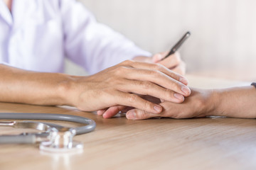 male doctor holding hand and comforting patient in a hospital with blur stethoscope in foreground on desk 