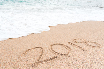 Happy new year 2018 concept. Number 2018 written on sandy beach with blue sea waves and foam.