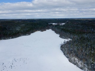 Aerial view during Winter over frozen lake (Vesslarpssjön) covered in snow, wild forest in the background, located in Skåne county in Sweden