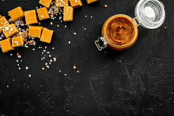 Contrast of flavors. The combination of salty and sweet. Caramel sauce in glass jar near caramel cubes on black background top view copy space