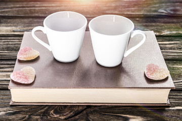 a pair of white mugs for coffee on a book on a wooden table. pleasant evening alone.