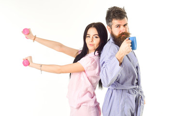 Couple in love in pajama, bathrobe stand isolated on white background. Couple, family on sleepy faces, full of energy. Girl with dumbbell, man with coffee cup. Morning alternative concept.