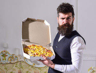 Pizza delivery concept. Man with beard and mustache holds delivered box with tasty fresh hot pizza. Macho in classic clothes hungry, on strict face, going to eat pizza, luxury interior background.