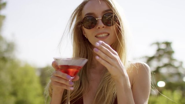 beautiful blonde with long hair smiling, showing off and flirting straightening hair, air kiss and turning in brown swimsuit and bikini with wineglass of cocktail a background of nature
