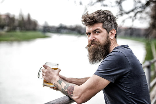 Side view bearded man enjoying big mug of beer on warm evening. Brutal man with messy hairstyle and tattooed arm having a drink in bar on the river font. Friday night after work