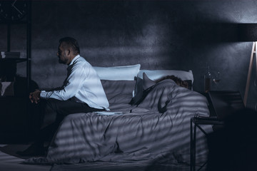 Dark gray room with a side view of a mature man sitting on a bed and having second thoughts after cheating and a woman sleeping under the cover