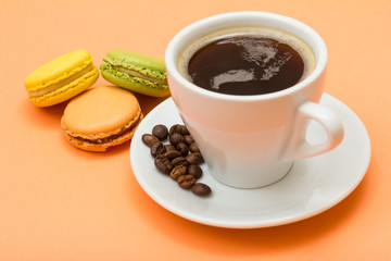 Cup of coffee with coffee beans and delicious macarons cakes of different color on peach background