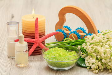 Obraz na płótnie Canvas Bottles with cream for face skin and aromatic oil, glass bowl with sea salt, burning candle, starfish, hand massager and bouquet of lilies of the valley