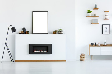 Black lamp next to fireplace in white living room interior with mockup of empty poster. Real photo
