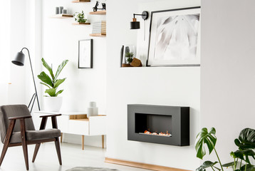 Grey wooden armchair in front of fireplace in white flat interior with poster and plant. Real photo