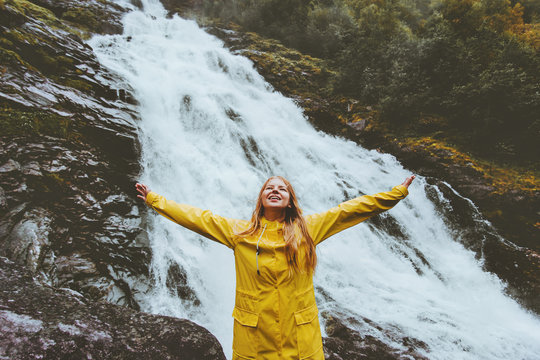 Happy smiling woman tourist raised hands enjoying waterfall landscape Traveling adventure lifestyle traveler wearing yellow raincoat success raised hands harmony with nature euphoria emotions
