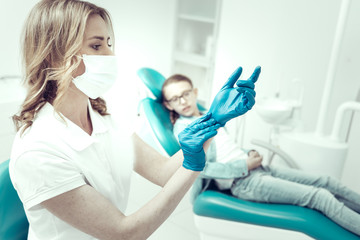 Necessary preparations. Serious concentrated young dentist putting on blue gloves before examining the patient