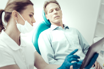 Detailed explanation. Serious concentrated patient listening to the recommendations of his dentist while sitting in a dental chair and looking at the screen of a tablet