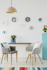 Creative living room interior with plates on the wall, table, grey armchairs, flowers and colorful rug
