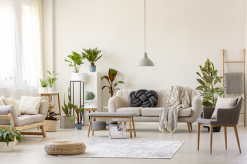 Pouf on rug and plants in spacious living room interior with grey chair near beige couch. Real photo - Powered by Adobe