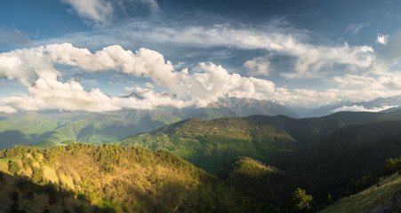 the tops of mountain ranges in the snow and covered by clouds in the summer on a sunny day with meadows and mountain slopes overgrown with trees, the Karachay-Cherkess Republic, the Caucasus