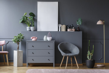 Grey armchair next to cabinet in flat interior with plants and mockup of white empty poster