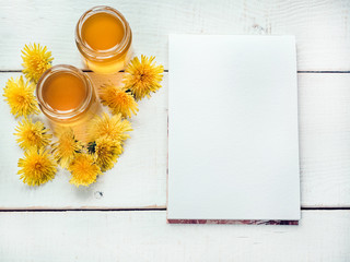 Yellow, bright dandelions, notebook with a blank page for Your inscriptions, flower honey in a jars on a beautiful, white, wooden table. Top view, close-up. Concept of a tasty and healthy meal