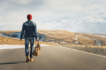 A stylish bearded hipster in sunglasses with a vintage backpack walks along the asphalt road on a sunny day. The concept of hitchhiking and hiking - obrazy, fototapety, plakaty