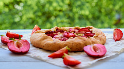 Homemade rhubarb tart on the gray kitchen table decorated with fresh sliced strawberries. Vegetarian healthy rhubarb galette on the blurred green background. Summer bakery dessert. Close up view