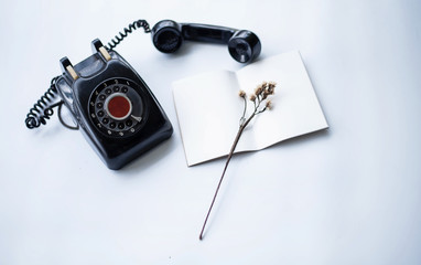 The antique telephone put beside the opened book with dried flower on white table,vintage style,blurry light around