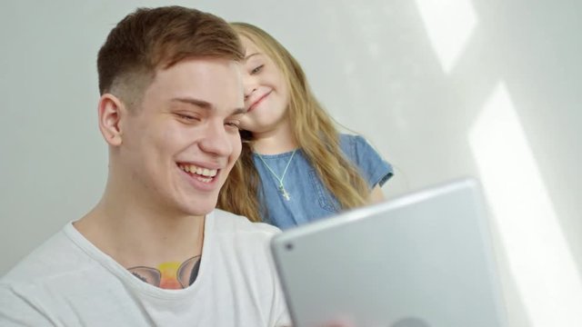 Medium shot of happy young father holding tablet and recording on frontal camera cute little girl making silly faces