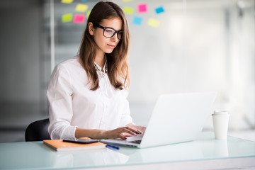 Portrait of beautiful smiling young brunette businesswoman sitting at bright modern work station and typing on laptop