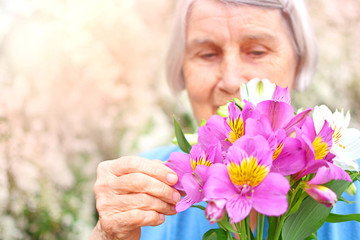 Happy old age concept. Beautiful aged woman looks at a bouquet of flowers in garden of her habitaion. Nursing home background..