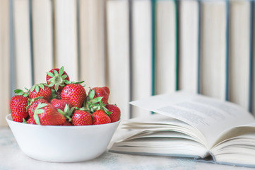 Fresh summer red strawberries in a white cap and open book on desk on background with books. The concept of healthy food and education. Leisure and reading books