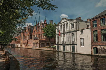 Obraz premium Wooded gardens and brick buildings on the canal's edge in a sunny day at Bruges. With many canals and old buildings, this graceful town is a World Heritage Site of Unesco. Northwestern Belgium.