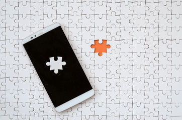 A modern big smartphone with several puzzle elements on the touch screen lies on a white jigsaw...